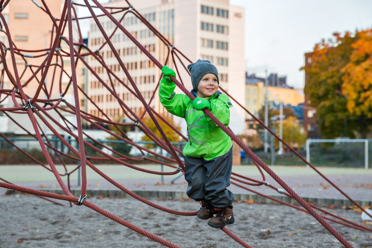 Kuvassa on lapsi kiipeämässä kiipeilytelineessä, taustalla näkyy taloja ja syksyisiä puita.